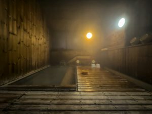 Steam filling a wooden indoor onsen bath with wooden buckets on a shelf