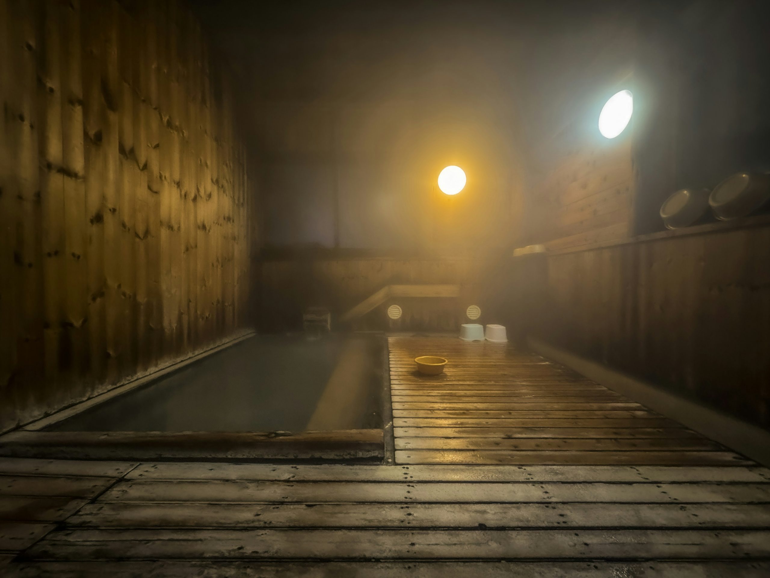Steam filling a wooden indoor onsen bath with wooden buckets on a shelf